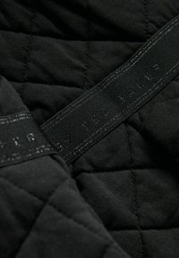 Tissu noir matelassé présentant une surface texturée. Une étiquette noire mate avec un lettrage en relief est visible, indiquant la marque.