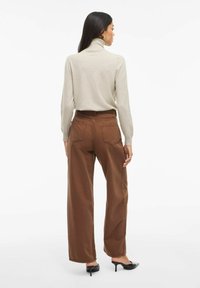 Maglione a collo alto beige chiaro, pantaloni larghi marroni con due tasche posteriori e mules nere con tacco. Tessuto morbido e vestibilità rilassata.