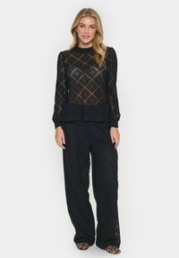 Top in pizzo nero con maniche lunghe caratterizzato da un motivo a diamante, abbinato a pantaloni neri a gamba larga. Entrambi gli articoli hanno una texture morbida.