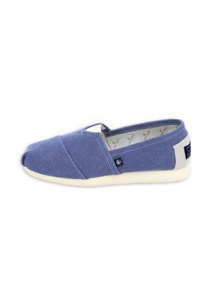 Zapato casual sin cordones con parte superior de tela azul, suela blanca y pequeñas etiquetas de tela en el lateral y el talón, mostrado desde una vista lateral.