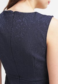 Marineblauwe jurk met een getextureerd bloemenpatroon, mouwloos ontwerp en een rits aan de achterkant. Gladde stof met een aangesloten silhouet.