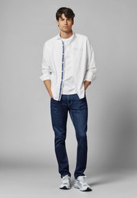Wit overhemd over een wit t-shirt, gecombineerd met donkerblauwe spijkerbroek en grijze sportschoenen. Model staat met handen in de zakken.