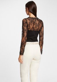 Zwarte kanten top met lange mouwen en ingewikkelde bloemenpatronen, gecombineerd met een strakke zwarte body eronder. Crèmekleurige broeken met een lichte glans.