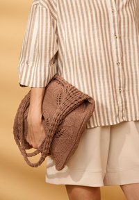 Personne portant une chemise rayée beige et blanche et un short crème, tenant un grand sac fourre-tout tricoté marron avec des poignées tressées.