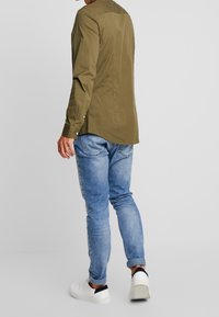 Man i olivgrön långärmad skjorta, ljusblå uppvikta jeans och vita sneakers, som står mot en enfärgad ljusgrå bakgrund.