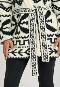 Svart och vit stickad cardigan med ett intrikat geometriskt mönster, vida ärmar och ett knytbälte med en dekorerad kant.