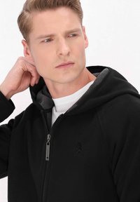 Svart zip-up hoodie i mjukt fleece-material, grå huva på insidan, dragkedja och diskret logotyp på bröstet.