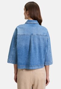 Giacca in denim azzurro chiaro con un design corto, cuciture visibili, un pieghe sul retro e maniche a tre quarti. Presenta un colletto classico.