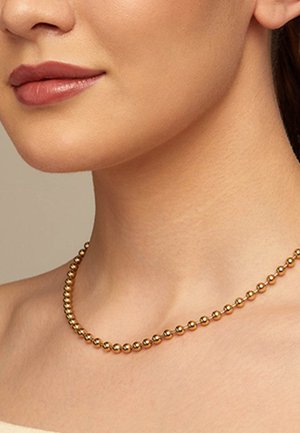 Primer plano del cuello de una mujer que lleva un collar dorado con cuentas lisas y redondas, con el tono natural de piel visible.