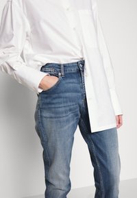 Camisa blanca de manga larga con botones y un bolsillo en el pecho, combinada con unos jeans azul medio, ligeramente desgastados, que presentan un diseño clásico de cinco bolsillos.