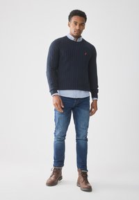 Pullover en maille torsadée dans un ton marine avec des poignets côtelés, superposé à une chemise à col bleu clair. Associé à un jean bleu et des bottes à lacets marron.