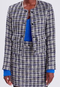 Tweed jacket in black, white, and blue plaid pattern, featuring a collarless design, front pockets, and black button closures. Blue sweater underneath.