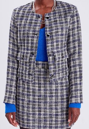 Veste en tweed à motif tartan noir, blanc et bleu, avec un design sans col, des poches avant et des boutons noirs. Un pull bleu en dessous.