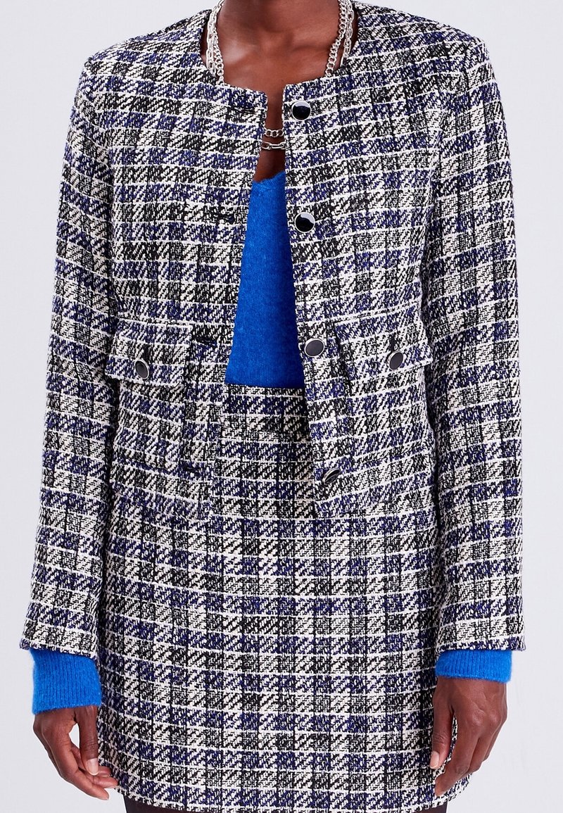 Tweed jacket in black, white, and blue plaid pattern, featuring a collarless design, front pockets, and black button closures. Blue sweater underneath.