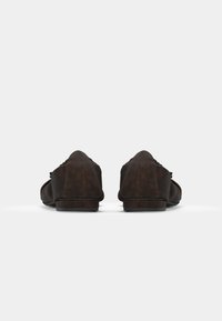 Mocassins en suède marron avec un petit talon, présentant un bout arrondi et un motif de couture décoratif sur le dessus. Texture lisse et design simple.