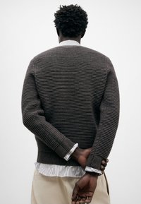 Suéter marrón de canalé con un diseño texturizado, combinado con una camisa a rayas visible en los puños y el dobladillo. Fondo neutro.