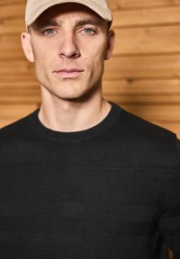 Pull noir en maille avec des rayures horizontales texturées. Le modèle porte une casquette beige, sur un fond en bois.