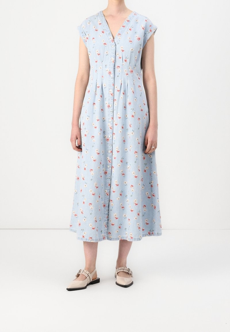 Femme portant une robe midi sans manches bleu clair avec un petit imprimé floral rouge et blanc et des ballerines beiges à bout pointu avec des brides à la cheville.