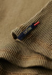 Brązowa prążkowana tkanina o tkanym wzorze, z niebieską metką naszytą z napisem "Superdry" oraz czerwonym akcentem nitki.