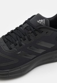 Primo piano di una sneaker Adidas nera con texture in rete, lacci neri e tre strisce parallele sul lato.