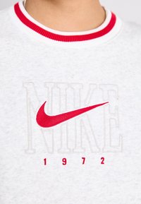 Felpa bianca con collo a costine rosso e bianco. Presenta il logo "Nike" ricamato in rosso e "1972" in grigio. Materiale morbido con una texture.