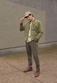 Giacca trapuntata verde oliva, camicia beige chiaro con bottoni, pantaloni oliva e scarpe marroni. Persona che sistema un cappellino beige; sfondo in cemento.