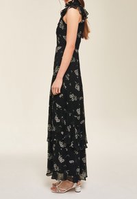 Svart blommig maxiklänning med volangdetaljer på axlarna, lager-på-lager design, halvgenomskinligt tyg och mönster av små vita och gröna blommor.