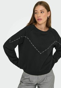 Femme portant un sweat-shirt noir avec des clous argentés en motif zigzag, un pantalon gris et des boucles d'oreilles créoles, regardant sur le côté avec les mains sur les hanches.