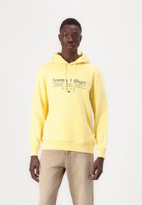Gelbe Kapuzenjacke aus weichem Stoff. Verfügt über eine Front-Kängurutasche, Kordeln und einen bedruckten Schriftzug auf der Brust. Ein Paar hellbraune Hosen.