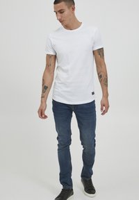Hvid bomulds T-shirt med korte ærmer, parret med blå denim jeans. Personen står foran en neutral baggrund iført sorte sneakers.