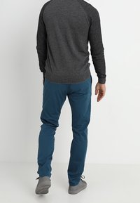Homme portant une chemise à manches longues gris foncé, un pantalon bleu et des chaussures grises, debout contre un fond clair uni, de dos.