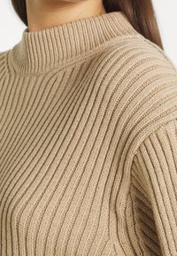 Beżowy sweter dziany z wysokim, prążkowanym kołnierzem i teksturą pionowych pasków. Charakteryzuje się luźnym krojem i miękką, średniej grubości tkaniną.