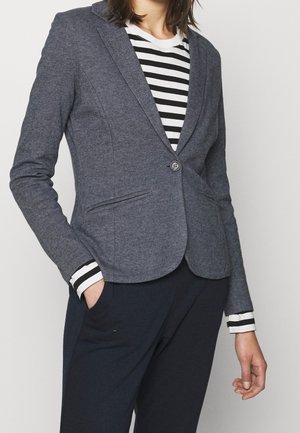 Femme portant un blazer gris à un bouton sur une chemise à manches longues rayée noir et blanc, avec un pantalon foncé et une main dans la poche.
