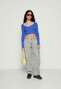 Kvinne iført en blå langermet cropped topp, lysegrå cargobukser, sorte joggesko, røde solbriller, holder en liten gul håndveske.