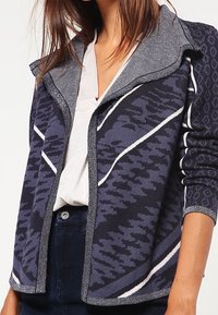 Stickad jacka med grå och marinblå mönstrad design, utrustad med en bred krage och vita accentlinjer. Mjuk struktur och öppen frontstil.