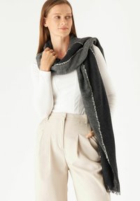 Écharpe grise au toucher doux, avec un bord effiloché et un discret motif. Portée légèrement sur un haut blanc et un pantalon beige.