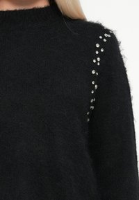 Melns adīts džemperis ar mīkstu, pūkainu tekstūru, kas papildināts ar izkaisītiem sudraba rhinestone akmeņiem pa vienu plecu, lai pievienotu sīku detaļu.
