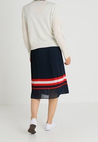 Maglione a maniche lunghe color beige chiaro abbinato a una gonna plissè blu navy con righe rosse e bianche. Sneakers bianche completano l'outfit.