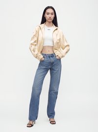 Femme portant une veste bomber en satin crème, un crop top blanc, un jean taille haute bleu et des talons ouverts noirs, debout les mains dans les poches.