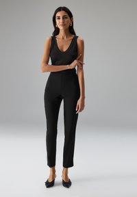 Vrouw in een zwarte mouwloze ribbeltop, slanke zwarte high-waist broek en spitse zwarte hakken, staand met gekruiste armen tegen een grijze achtergrond.