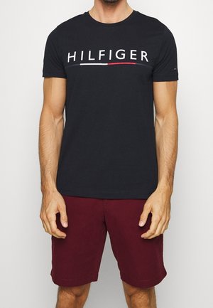 Mand iført sort Hilfiger t-shirt med hvidt og rødt logo og bordeaux shorts stående mod en hvid baggrund.