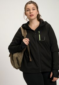 Giacca nera con zip e cappuccio, con un accento verde sulla zip, tessuto testurizzato e una piccola tasca sul petto. Porta uno zaino beige.