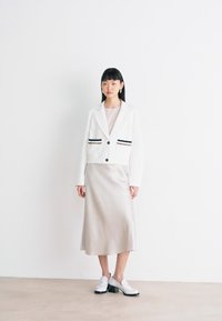 Valkoinen lyhyt bleiseri mustilla ja beige-raidoilla, päällä beige midi-mekko. Malli seisoo valkoisessa tilassa, jalassa valkoiset kengät.