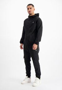 Zwarte sportjack met aansluitende capuchon, ritssluiting en bijpassende zwarte cargobroek. Kenmerken zijn elastische manchetten en zijzakken. Witte sneakers.