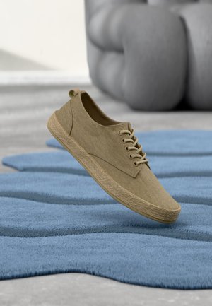Beige casual lace-up shoe floating above a blue textured rug with blurred sofa in the background.