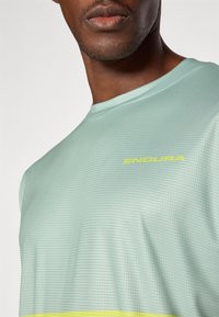 Maglietta sportiva verde chiaro con una texture a griglia, con un logo giallo "ENDURA" e una striscia gialla orizzontale vicino all'orlo.