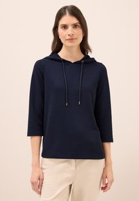 Pullover con cappuccio blu navy, texture a coste, maniche a tre quarti, cordino regolabile, vestibilità leggermente rilassata, abbinato a pantaloni beige chiari.