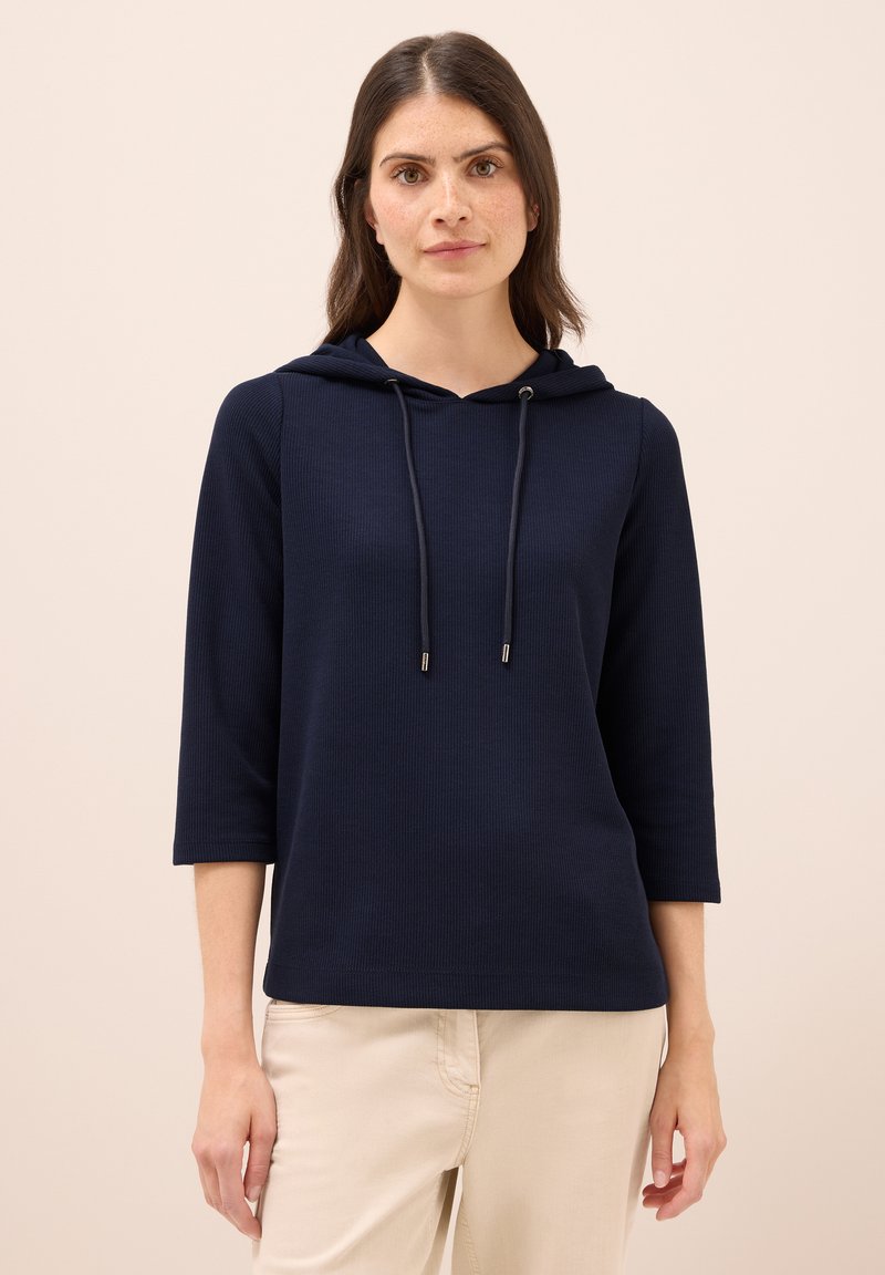 Pullover con cappuccio blu navy, texture a coste, maniche a tre quarti, cordino regolabile, vestibilità leggermente rilassata, abbinato a pantaloni beige chiari.