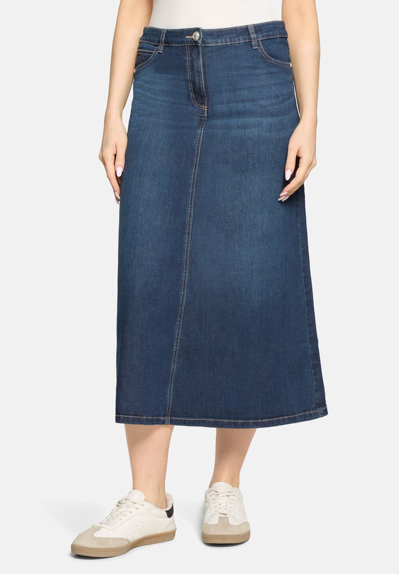 Kvinde iført en midilengde mørkeblå denimnederdel med fremadgående syninger og hvide sneakers på hvid baggrund.