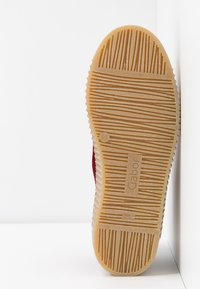 Semelle de chaussure en caoutchouc avec un motif de bande de roulement et le nom de la marque "Gabor", positionnée verticalement contre un coin blanc en arrière-plan.